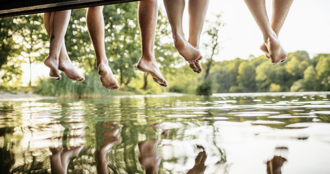 Man ser benen och fötterna på unga som sitter på en brygga vid en sjö