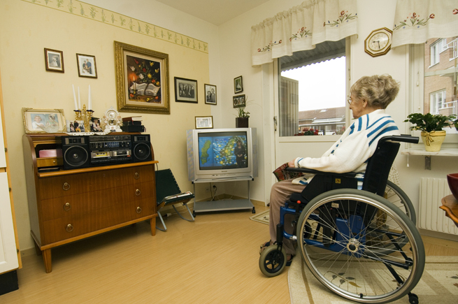 Bild på en äldre dam som sitter i en rullstol och kollar på tv