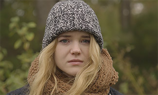 Bild på en ung kvinna som står utomhus med grå mössa och tittar in i kameran med ett bekymrat ansiktsuttryck