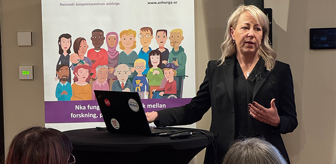 En kvinna som står vid ett bord och föreläser framför en publik 