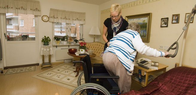 Bild på en kvinna som hjälper en äldre kvinna att sätta sig i rullstolen