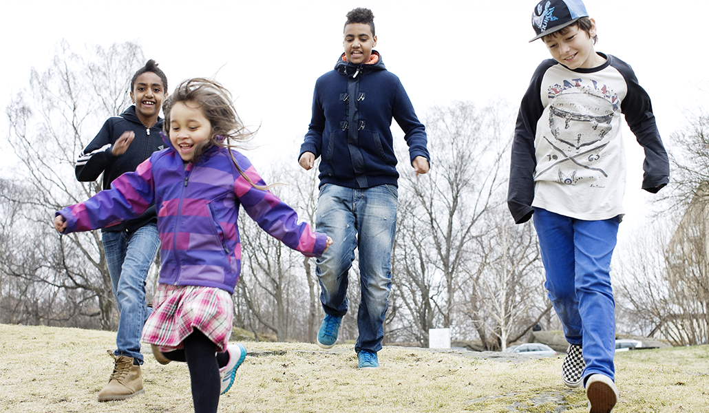 Några barn springer och hoppar på en gräsmatta. De är glada. 