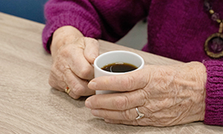 En äldre kvinna sitter vid ett bord och håller i en kopp med kaffe.