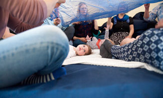 Bebisar ligger på golvet med vuxna runt omkring som håller i ett blått genomskinligt tyg.