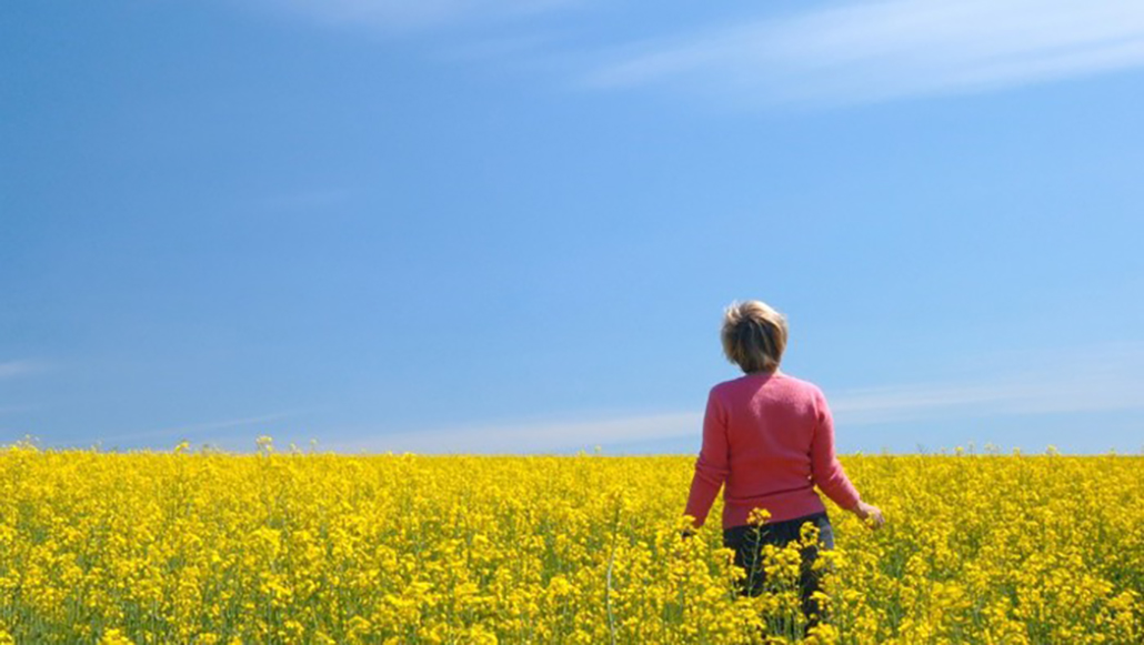 En kvinna går i ett gult rapsfält. Himlen är klarblå med några få moln.