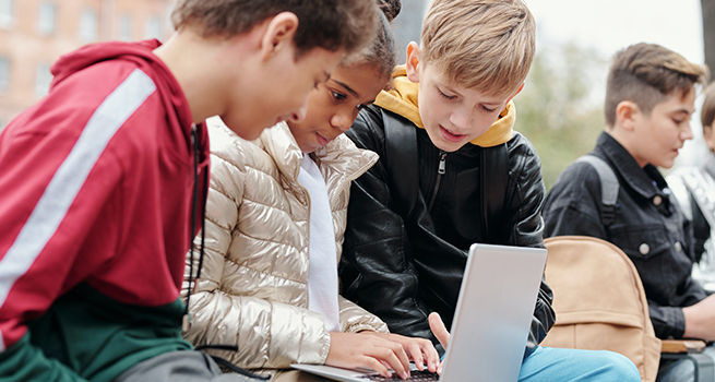Elever sitter på rad. En flicka i mitten har en dator i knät och två pojkar sitter bredvid och tittar på datorn.