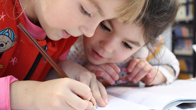 Två barn lutar sig över ett ritblock. Det ena barnet håller i en penna och ritar.