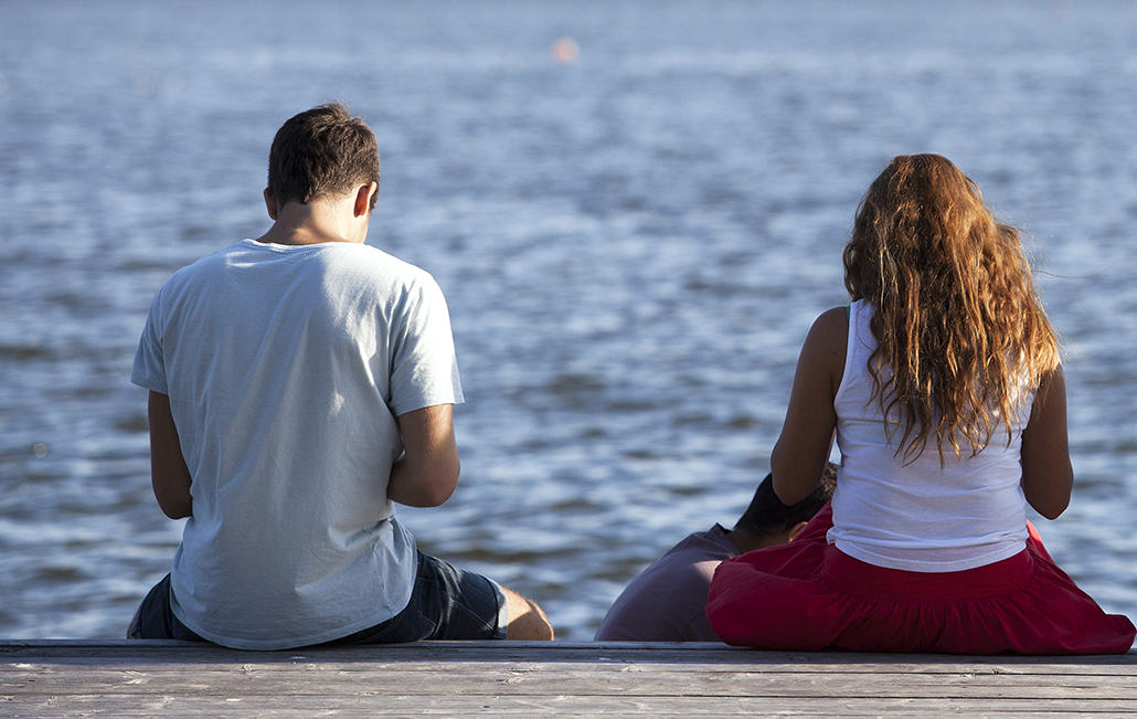 Två personer sitter på en brygga vid vattnet med ryggen mot kameran.
