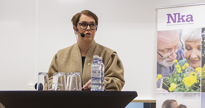 Socialminister Lena Hallengren står vid ett ståbord och talar.