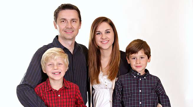 En familj bestående av en mamma, pappa och två söner står framför en vit vägg och tittar in i kameran.